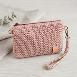 May include: A dusty rose crochet zippered pouch featuring a rectangular shape, a top metal zipper closure, and a matching braided wrist strap attached to a brass-toned metal clasp. A small rectangular leather-like tab is sewn on the bottom right.