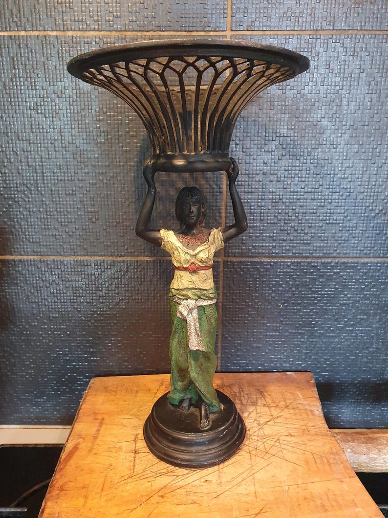 May include: A decorative stand featuring a dark-toned figure standing on a circular base, holding a large, black, open-work metal bowl above their head. The figure wears a yellow top with a red waist detail and a long green skirt with a white sash.