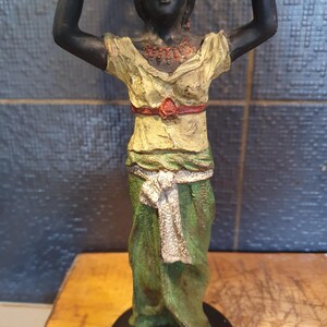 May include: A standing figurine of a person with dark skin, wearing a pale yellow top with a red waist belt and a green skirt with a white sash. The figure has arms raised above the head, a red necklace, and is mounted on a circular black base.