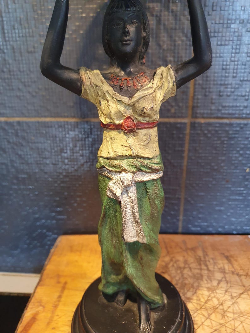 May include: A standing figurine of a person with dark skin, wearing a pale yellow top with a red waist belt and a green skirt with a white sash. The figure has arms raised above the head, a red necklace, and is mounted on a circular black base.