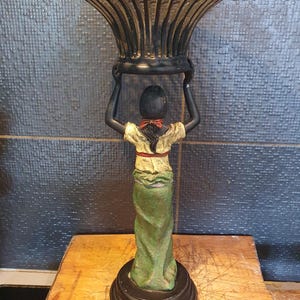 May include: A decorative stand featuring a sculpted figure with dark skin, wearing a cream-colored top with a red sash and a long green skirt. The figure holds a black, wide-mouthed, lattice-patterned metal bowl above their head on a circular base.