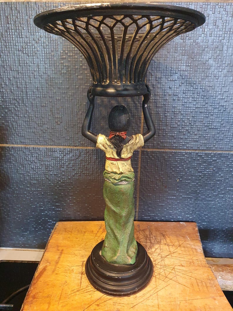 May include: A decorative stand featuring a sculpted figure with dark skin, wearing a cream-colored top with a red sash and a long green skirt. The figure holds a black, wide-mouthed, lattice-patterned metal bowl above their head on a circular base.