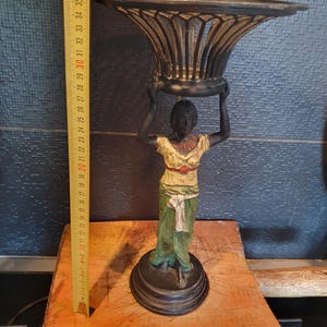May include: A black metal decorative stand featuring a figure holding a bowl overhead. The figure wears a cream-colored top with a red accent and a green skirt with a white sash. A yellow measuring tape shows the object is approximately 15 inches tall.