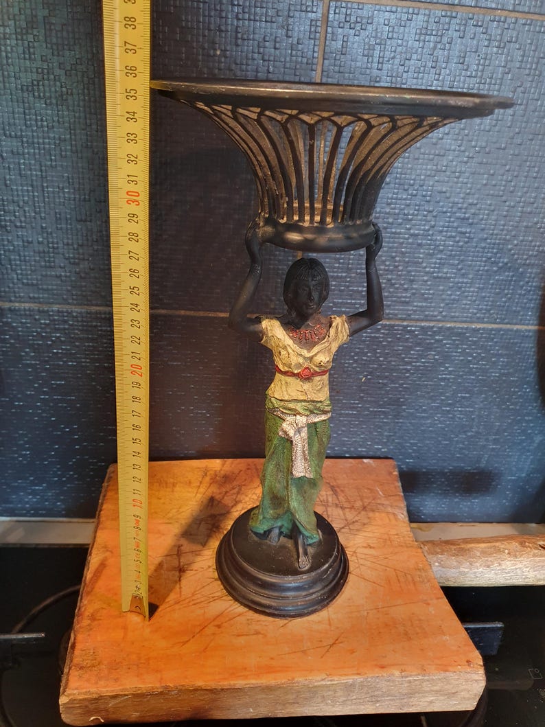 May include: A black metal decorative stand featuring a figure holding a bowl overhead. The figure wears a cream-colored top with a red accent and a green skirt with a white sash. A yellow measuring tape shows the object is approximately 15 inches tall.
