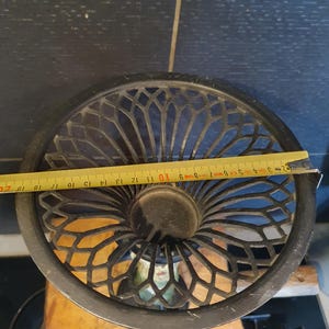May include: A black metal candle holder with a wide, circular, lattice-patterned top. A yellow measuring tape is stretched across the diameter, showing a width of approximately 9.5 inches. The structure features a central circular opening.