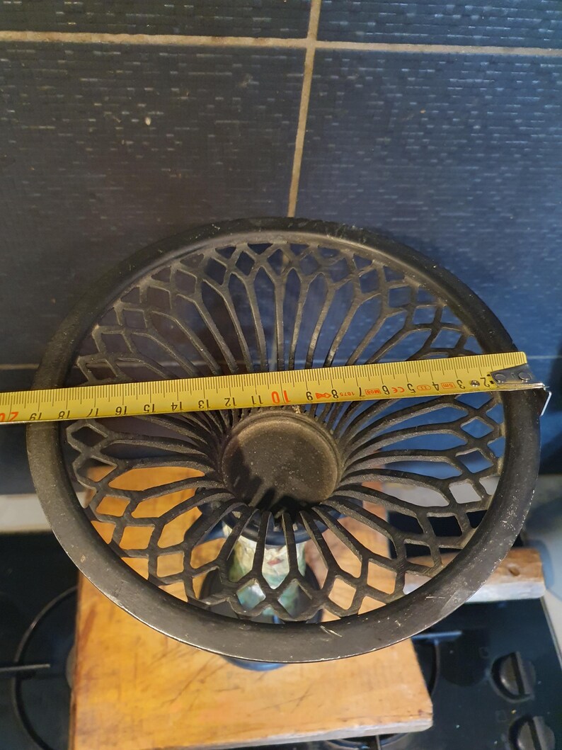 May include: A black metal candle holder with a wide, circular, lattice-patterned top. A yellow measuring tape is stretched across the diameter, showing a width of approximately 9.5 inches. The structure features a central circular opening.