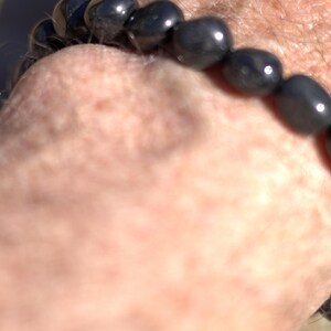 May include: A dark gray beaded bracelet with oval-shaped beads. The bracelet is worn on a wrist, with the beads reflecting light. The bracelet appears to be made of polished stone or a similar material. The bracelet is a piece of jewelry.