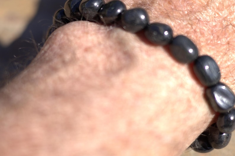 May include: A dark gray beaded bracelet with oval-shaped beads. The bracelet is worn on a wrist, with the beads reflecting light. The bracelet appears to be made of polished stone or a similar material. The bracelet is a piece of jewelry.