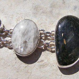 Pode incluir: Um bracelete de corrente de prata com quatro pingentes de pedras preciosas diferentes. Os pingentes são uma pedra branca, uma pedra verde e branca, uma pedra preta e verde e uma pedra branca.