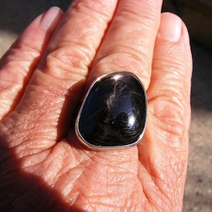 Peut inclure: Une bague en argent avec une grande pierre précieuse noire de forme ovale. La pierre précieuse a une surface réfléchissante et semble avoir un motif en spirale.