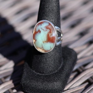 May include: Oval gemstone ring with a silver band. The stone features a blend of light green and reddish-brown hues. The ring is displayed on a black ring holder, showcasing its design and colors. A unique piece of jewelry.