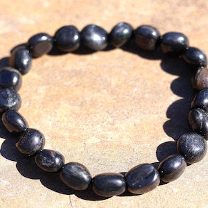 May include: A black beaded bracelet made of polished, oval-shaped stones. The bracelet is strung together, forming a circular shape. The stones have a glossy finish and reflect light, creating highlights.