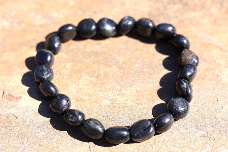 May include: A black beaded bracelet made of polished, oval-shaped stones. The bracelet is strung together, forming a circular shape. The stones have a glossy finish and reflect light, creating highlights.
