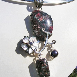 May include: A silver pendant necklace featuring a red and green gemstone, a white flower with a black centre, and a black gemstone dangle. The pendant is attached to a silver chain.