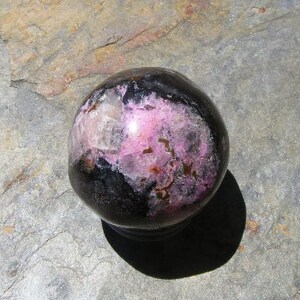 May include: A polished sphere of black and pink rhodochrosite, a type of mineral, with a smooth, shiny surface.