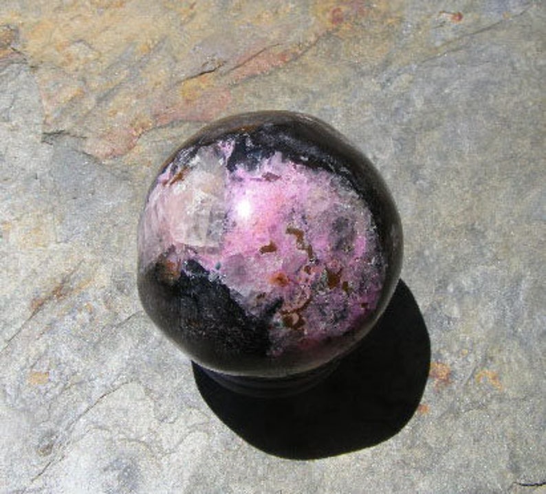 May include: A polished sphere of black and pink rhodochrosite, a type of mineral, with a smooth, shiny surface.