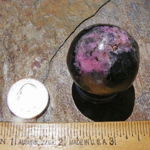 May include: A polished black and pink sphere with a smooth, shiny surface. The sphere is approximately 2 inches in diameter and is resting on a brown, textured surface.