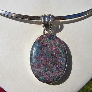 Puede incluir: Un collar de plata con un gran colgante ovalado con una piedra preciosa verde y rosa. El colgante está engastado en plata y tiene un diseño detallado.