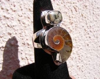 May include: A silver ring with a large ammonite fossil and a smaller, square, gray and white stone. The ring is on a black velvet display stand.