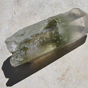 Puede incluir: Un gran cristal de cuarzo transparente con una inclusión verde y musgosa. El cristal está parcialmente cubierto de una sustancia blanca y pulverulenta.