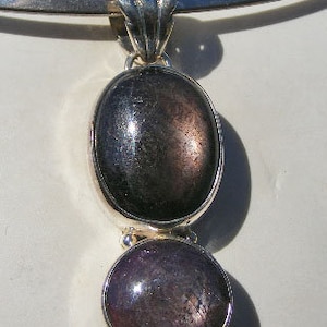 May include: A silver pendant with two oval-shaped gemstones. The larger gemstone is a dark gray color with a shimmery finish. The smaller gemstone is a light purple color with a shimmery finish.