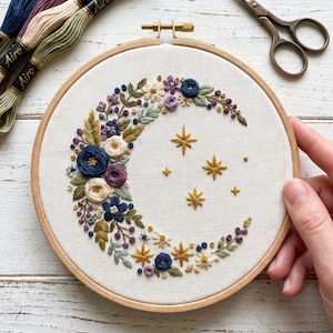 Puede incluir: Un bastidor de bordado circular de madera que contiene tela blanca con un diseño de media luna bordado. La luna está formada por flores azules, crema y moradas con hojas verdes, acentuada por varias estrellas amarillas bordadas.