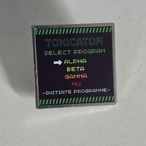 Op de afbeelding: Een vierkante metalen speld met een zwarte achtergrond en zilveren rand. De tekst luidt TOXICATOR, SELECT PROGRAM, ALPHA, BETA, GAMMA, MU en -INITIATE PROGRAMME-. Groene diagonale strepen sieren de boven- en onderkant. Een witte pijl wijst naar ALPHA.