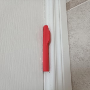 Könnte beinhalten: Eine leuchtend rote, längliche, rechteckige Türscharnierabdeckung aus Kunststoff mit einer kleinen hervorstehenden Lasche an der Seite. Die Abdeckung ist vertikal am Rand eines weißen Türrahmens angebracht und hat eine glatte, matte Oberfläche.