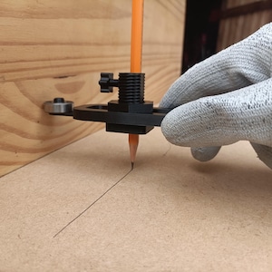 May include: A black scribing tool with a small metal wheel guide and a central holder for an orange pencil. The tool is positioned against a wooden surface to mark a straight line on a flat board, featuring a black adjustment knob.