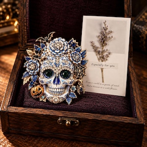 May include: A skull brooch featuring blue and white enamel, clear rhinestones, and green eye accents. The design includes floral motifs, a small bat, a spider, and a pumpkin. Beside it is a card with a dried plant and the text Especially for you.