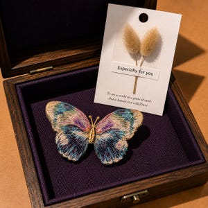 May include: A butterfly brooch with iridescent blue, purple, and gold embroidery, featuring a metallic gold body. It is presented in a dark wooden box alongside a white card with dried grass, the text Especially for you, and a poetic quote.