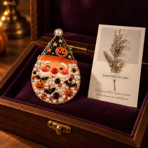 Puede incluir: Un broche en forma de lágrima con un rostro de Santa Claus de nariz roja, que lleva un sombrero de bruja negro con calabazas naranjas y estrellas. La barba está decorada con perlas blancas, cristales naranjas, cristales negros y murciélagos.