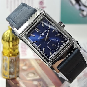 May include: A rectangular wristwatch with a polished silver-tone stainless steel case and a deep blue sunray dial. The dial features silver-tone hour markers, silver hands, and the Jaeger-LeCoultre logo. It has a dark blue leather strap.