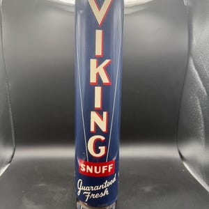 May include: A cylindrical blue metal container for Viking Snuff. The word VIKING is printed vertically in large white letters with red outlines. Below this, the word SNUFF appears in white on a red background, followed by the text Guaranteed Fresh.
