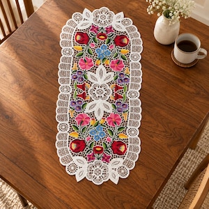 Könnte beinhalten: Ein ovaler weißer Spitzentischläufer mit aufwendigen Lochstickerei-Rändern und einem zentralen Blumenstickmuster in leuchtenden Farben wie Rot, Pink, Violett, Blau und Gelb, symmetrisch angeordnet mit grünen Blättern auf weißem Stoffgrund.