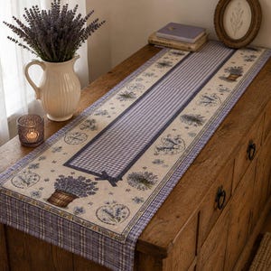 Puede incluir: Un camino de mesa rectangular con un panel central de cuadros vichy morados y blancos, bordeado por una tela color crema decorada con estampados de ramas de lavanda, macetas de lavanda y motivos florales en forma de corazón en tonos morados y verdes.