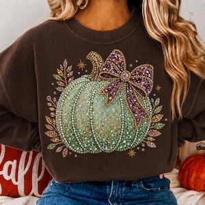 Könnte beinhalten: Ein dunkelbraunes Sweatshirt mit Rundhalsausschnitt und einem großen Motiv eines hellgrünen Kürbisses, der mit einer auffälligen, glitzernden violetten und rosafarbenen Schleife verziert ist. Der Kürbis ist von dekorativen, glitzernden Herbstblättern und kleinen goldenen Sternen umgeben.