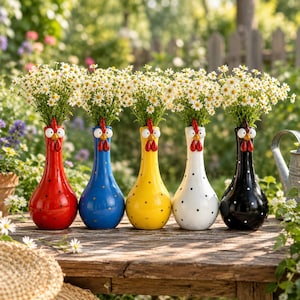 Op de afbeelding: Vijf keramische vazen in de vorm van kippen, elk met een lange hals, brede basis en stippenpatroon. De vazen zijn rood, blauw, geel, wit en zwart en elk gevuld met een boeket van kleine, witte, madeliefachtige bloemen.
