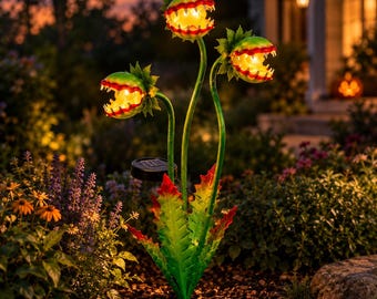 May include: A solar-powered garden stake featuring three green and red Venus flytrap-shaped heads with jagged teeth and glowing yellow centers. The plant has long, slender green stems and serrated leaves at the base, with a small black solar panel.