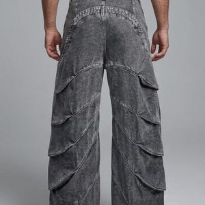 May include: Wide-leg denim trousers in a dark grey acid wash finish. The trousers feature a high-waisted design with multiple horizontal layered fabric panels running down the length of each leg, creating a tiered effect. The hem includes adjustable drawstrings.