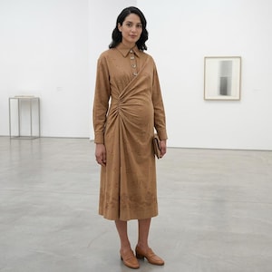 Puede incluir: Un vestido midi de color tostado con mangas largas, cuello camisero y cierre de botones frontal. El vestido presenta un detalle fruncido en la cintura y un sutil patrón floral bordado a lo largo del dobladillo, combinado con mocasines tostados.