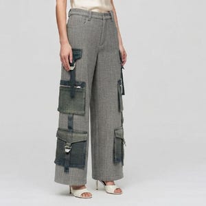 May include: Grey herringbone trousers featuring a wide-leg silhouette and multiple dark grey cargo pockets. The pockets are secured with zips, straps, and metallic buckles, creating a layered look on the front and sides of the garment.
