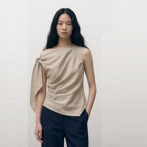 May include: A beige sleeveless top featuring an asymmetrical neckline and a knotted detail on one shoulder. The garment has a draped, gathered fabric construction that falls to the waist, paired with dark navy blue trousers with a straight leg.