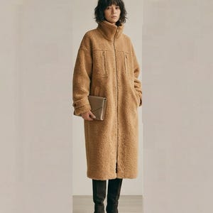 Könnte beinhalten: Ein langer, hellbrauner Teddy-Fleece-Mantel mit hohem Kragen, durchgehendem Frontreißverschluss und zwei vertikalen Reißverschlusstaschen auf der Brust. Der Mantel hat lange Ärmel mit geknöpften Manschetten und wird mit dunklen Stiefeln kombiniert.