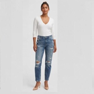 May include: A white long-sleeve ribbed top with a V-neckline paired with blue denim jeans featuring a distressed finish, ripped knees, and a classic five-pocket design. The jeans have a straight-leg cut and a medium wash with fading details.