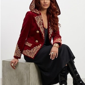 May include: A deep red velvet hooded jacket featuring intricate gold embroidery along the lapels, cuffs, and hem. The jacket has a double-breasted front with four gold-toned buttons, a structured collar, and a hood with matching gold trim.