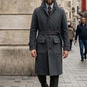 Puede incluir: Un abrigo de mezcla de lana gris carbón con solapa de muesca, cintura ceñida con hebilla rectangular, dos grandes bolsillos con solapa y puños abotonados. Se lleva sobre una camisa de cuello azul claro y una corbata de color marrón.