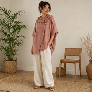May include: A dusty rose hooded poncho featuring a single button closure at the neckline, paired with loose-fitting off-white wide-leg trousers. The poncho has short sleeves and a draped silhouette, while the trousers reach the ankles.