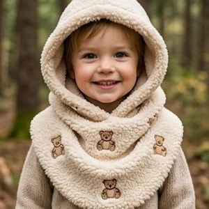 May include: A cream-colored, textured sherpa fleece hooded cowl featuring embroidered brown teddy bear motifs scattered across the fabric. The garment includes a deep, cozy hood and a wide, draped neck warmer section that covers the shoulders.