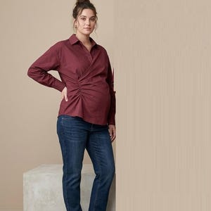 May include: A long-sleeved burgundy button-down shirt with a gathered side detail, paired with dark blue denim jeans and dark brown leather loafers. The shirt features a classic collar and a wrap-style front with ruched fabric at the waist.
